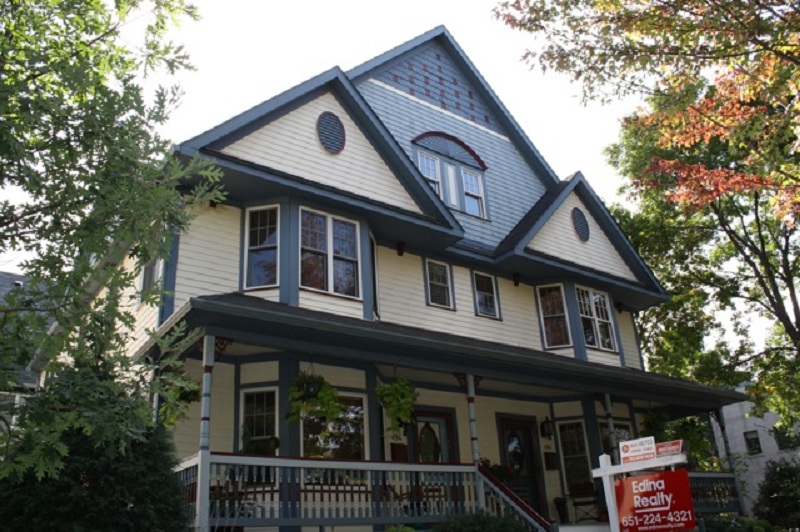 Victorian-style house with a mix of soft beige and blue siding, accented by greenery and a "For Sale" sign, showcasing modern curb appeal in a suburban Minnesota setting.