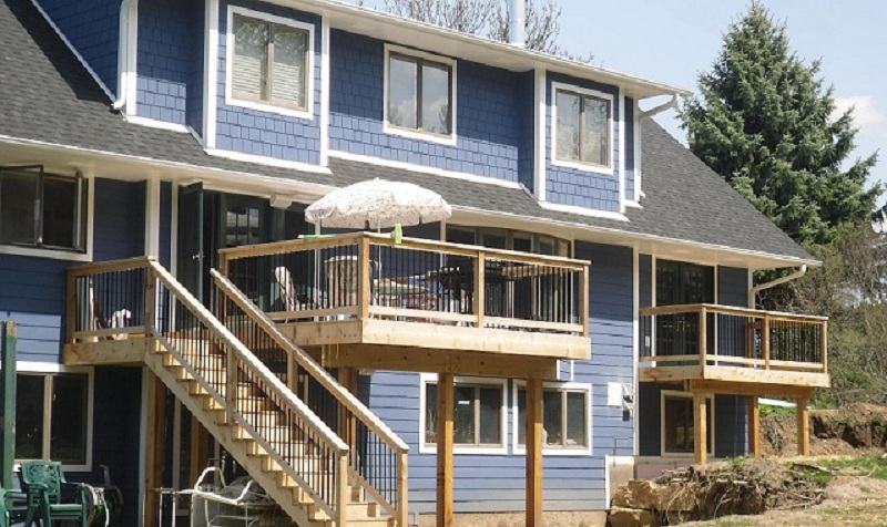 Blue two-story house featuring wooden decks with black railings, surrounded by greenery, showcasing modern siding design relevant to Minnesota's residential construction trends.